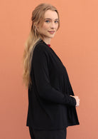 Woman stands side on in black soft, stretch rayon cardi with long sleeves, holding the front hem