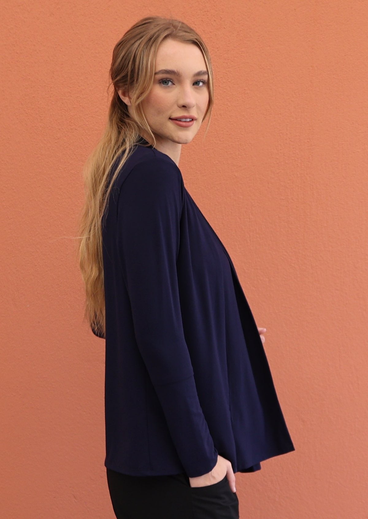 Woman standing sideways wears a full sleeved, soft rayon stretch cardigan in navy blue with a hand in pocket of pants.