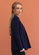 Woman standing sideways wears a full sleeved, soft rayon stretch cardigan in navy blue with a hand in pocket of pants.