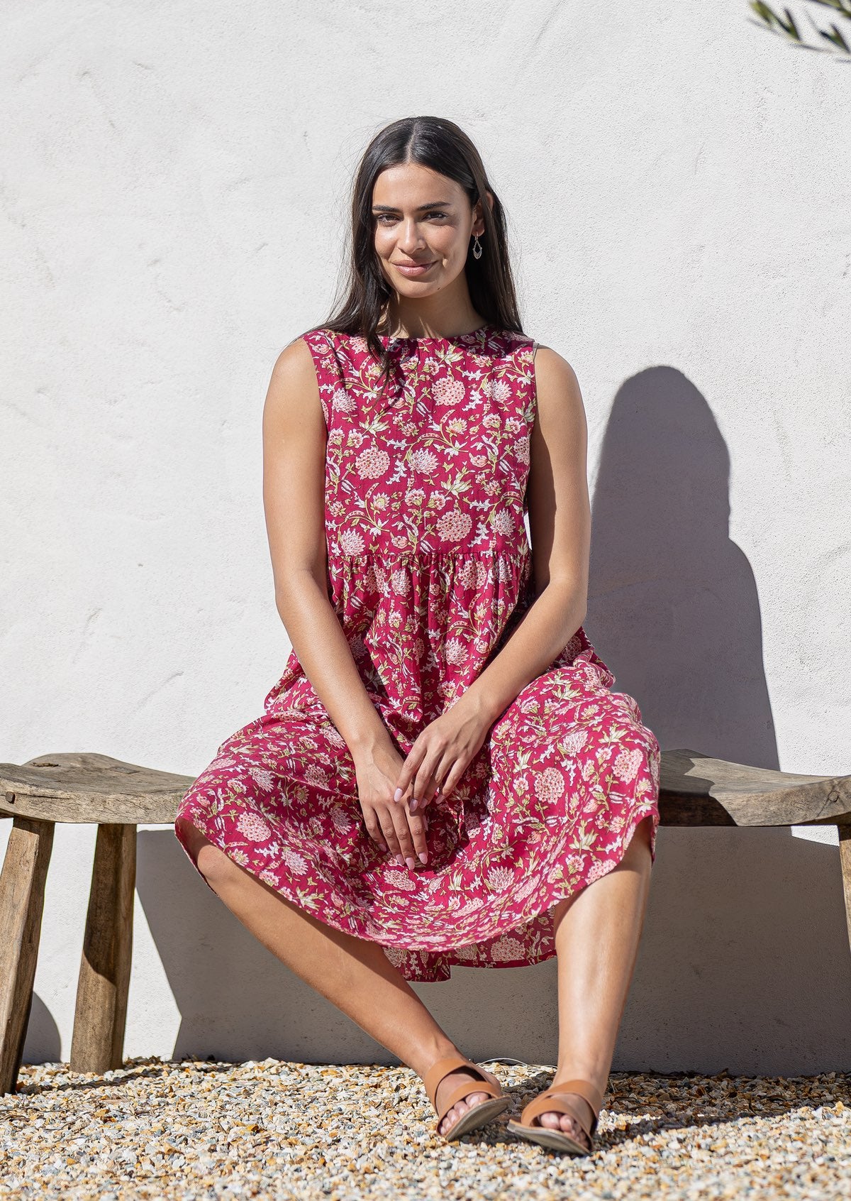 Woman in a pink floral cotton sundress sitting on a wooden bench against a white wall