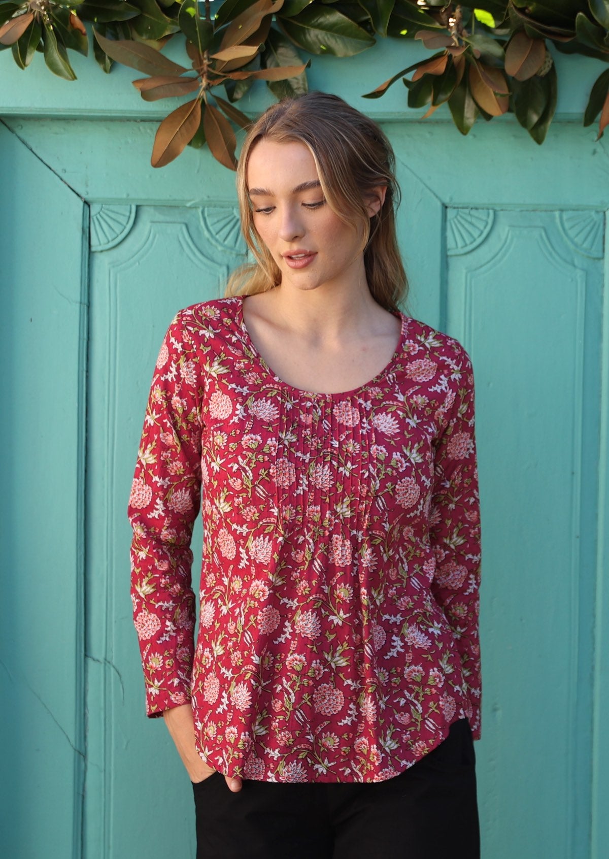 Model standing near aqua door wears lightweight cotton top with long sleeves and tiny pleats from the neckline.