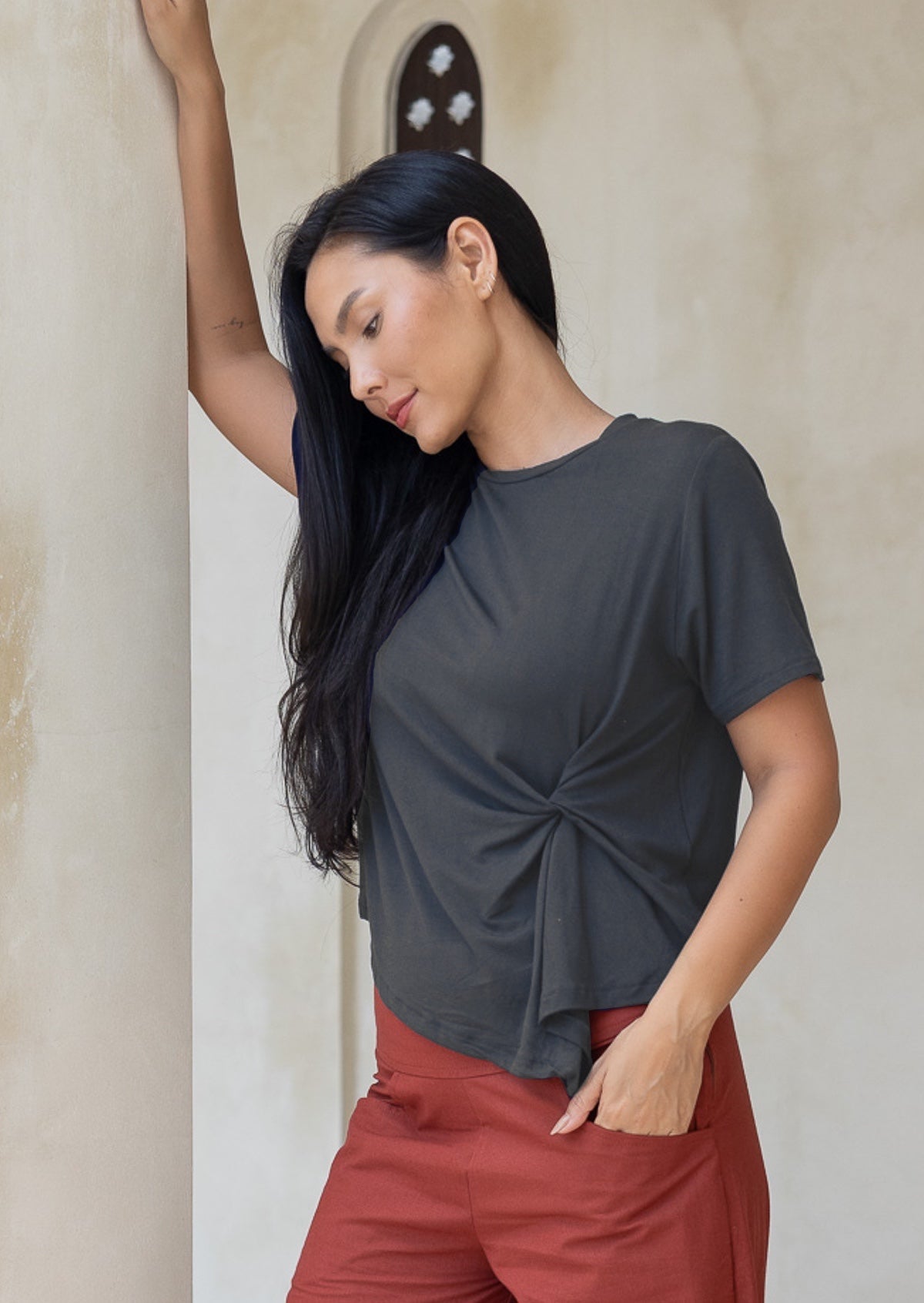 Woman leans against pillar wearing a dark grey rayon t-shirt with short sleeves and knot detail in the front, paired with rust coloured pants