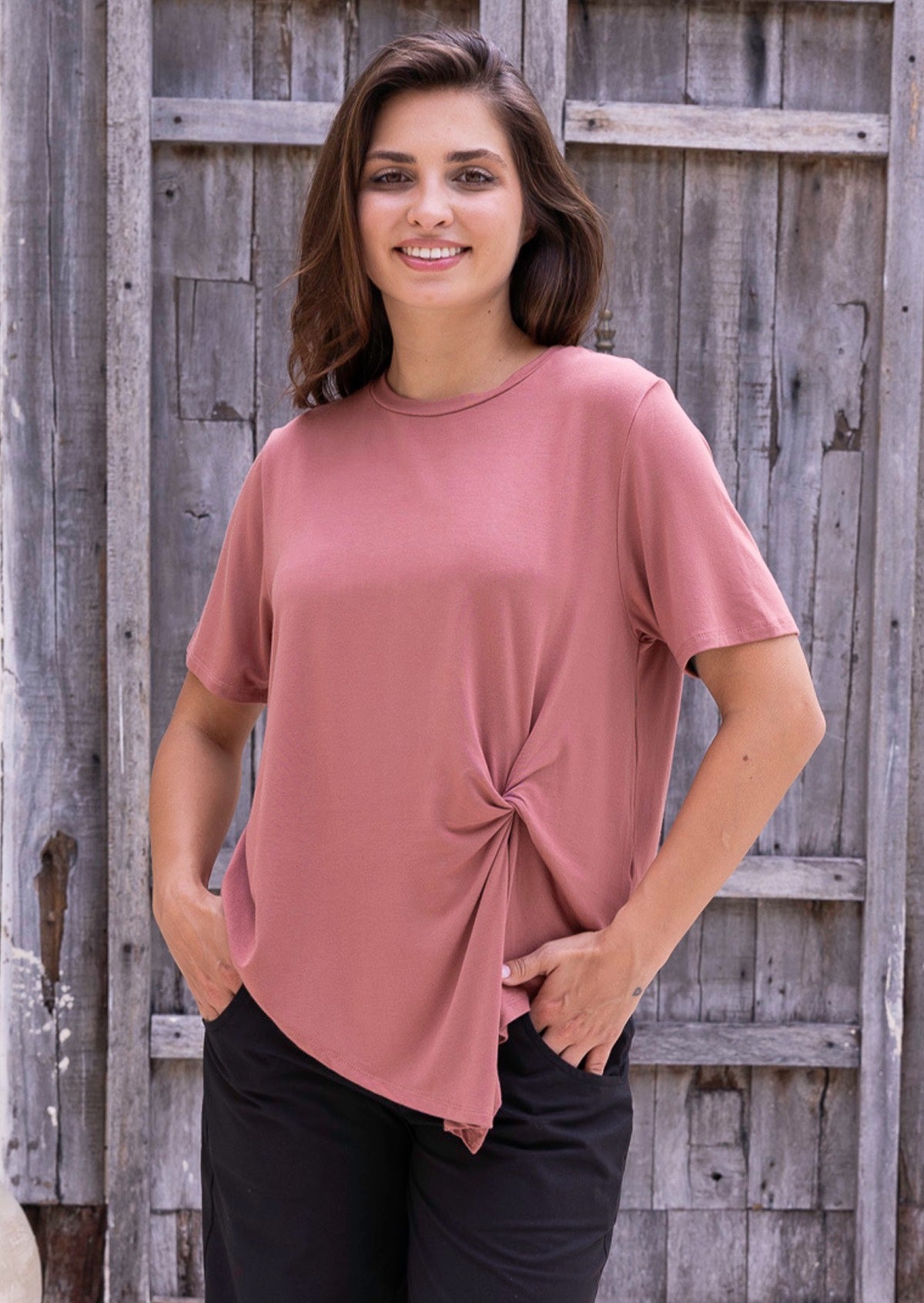 Woman wearing a pink t-shirt with a knot detail over black cotton pants with hands in pockets