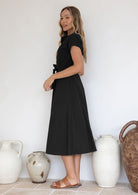 Woman wearing a black 50's style midi length dress with cap sleeves, standing in front of decorative vases.