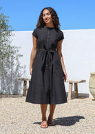 Woman wearing a black cotton retro style dress with button down front standing outdoors with a white wall with hands in pockets.