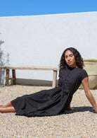 Woman in a black cotton dress sitting on pebbles with a white wall and blue sky in the background