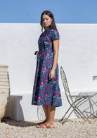 Woman wearing a cotton floral midi dress standing against a white wall with a blue sky.