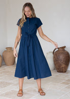 Model wears blue button through cotton dress with high round neckline, short sleeves and small pleats at center waist