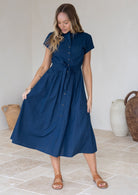 Woman wears cotton button through 50's style dress in plain deep blue shade with fabric waist tie, short sleeves and hidden side pockets