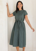 Woman wearing a moss green button through midi length dress with gathers at the waist, cinched with a waist tie