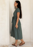 Woman standing side on wearing a green midi length dress with cap sleeves and a waist tie