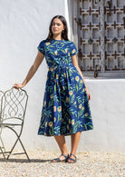 Woman wearing a blue floral cotton midi dress standing next to a chair against a white wall.