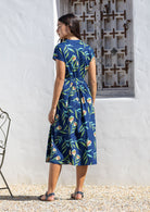 Woman wearing a blue floral cotton sundress standing in front of a white wall with a decorative metal gate.