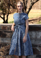 Woman leans on stone wall in blue based floral print cotton button through 50's style dress