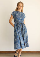 Woman wears cotton button through 50's style dress in floral print on a blue base, with hands in hidden side pockets