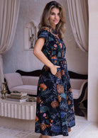 Model stands slightly side ways in 100 percent cotton retro button through dress with orange and pink florals; blue, green and grey leafy design on black base.