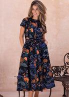 Model wearing a black floral cotton, button through dress with removable waist tie, short sleeves and hands in pocket.