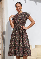 Woman wears black based floral printed cotton button through dress with hidden side pockets, short sleeves and a waist tie.