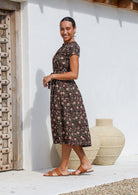 Women stands in side pose wearing floral printed midi length dress with high round neckline, short sleeves and pockets.