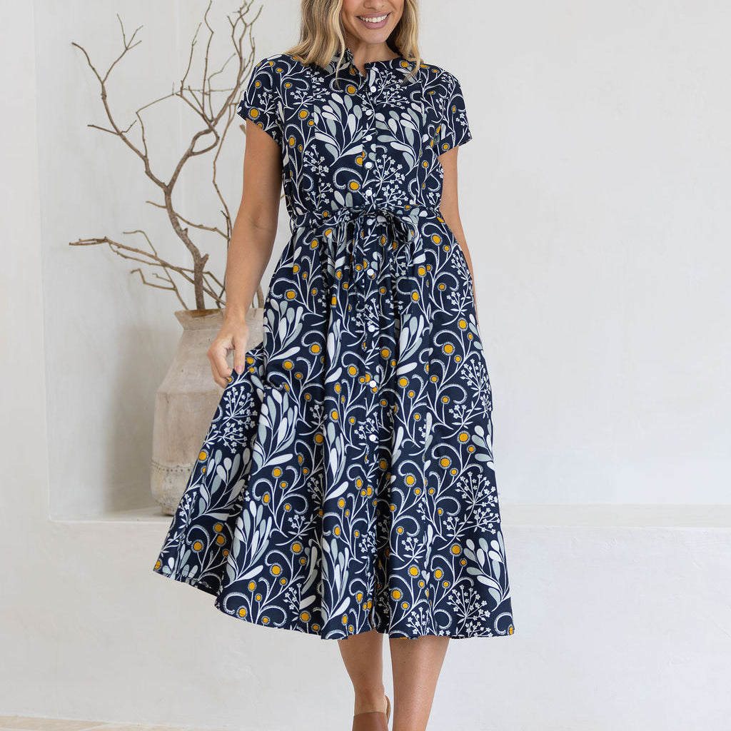 Woman wearing a dark cotton floral dress standing in a minimalistic room with a vase and branches.