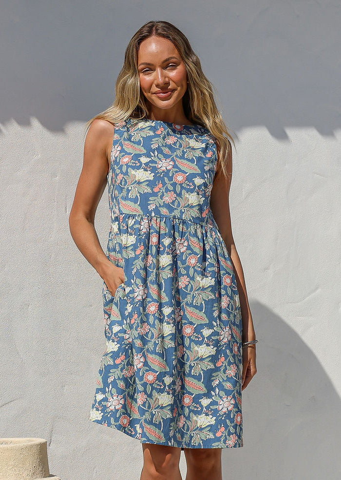 Woman in a above knee length 100% cotton floral dress standing against a white wall with a large beige pot nearby