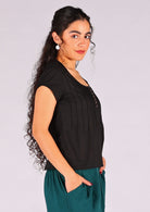 Model stands with her side to the front in black 100% cotton cap sleeve top with elastic in the trim