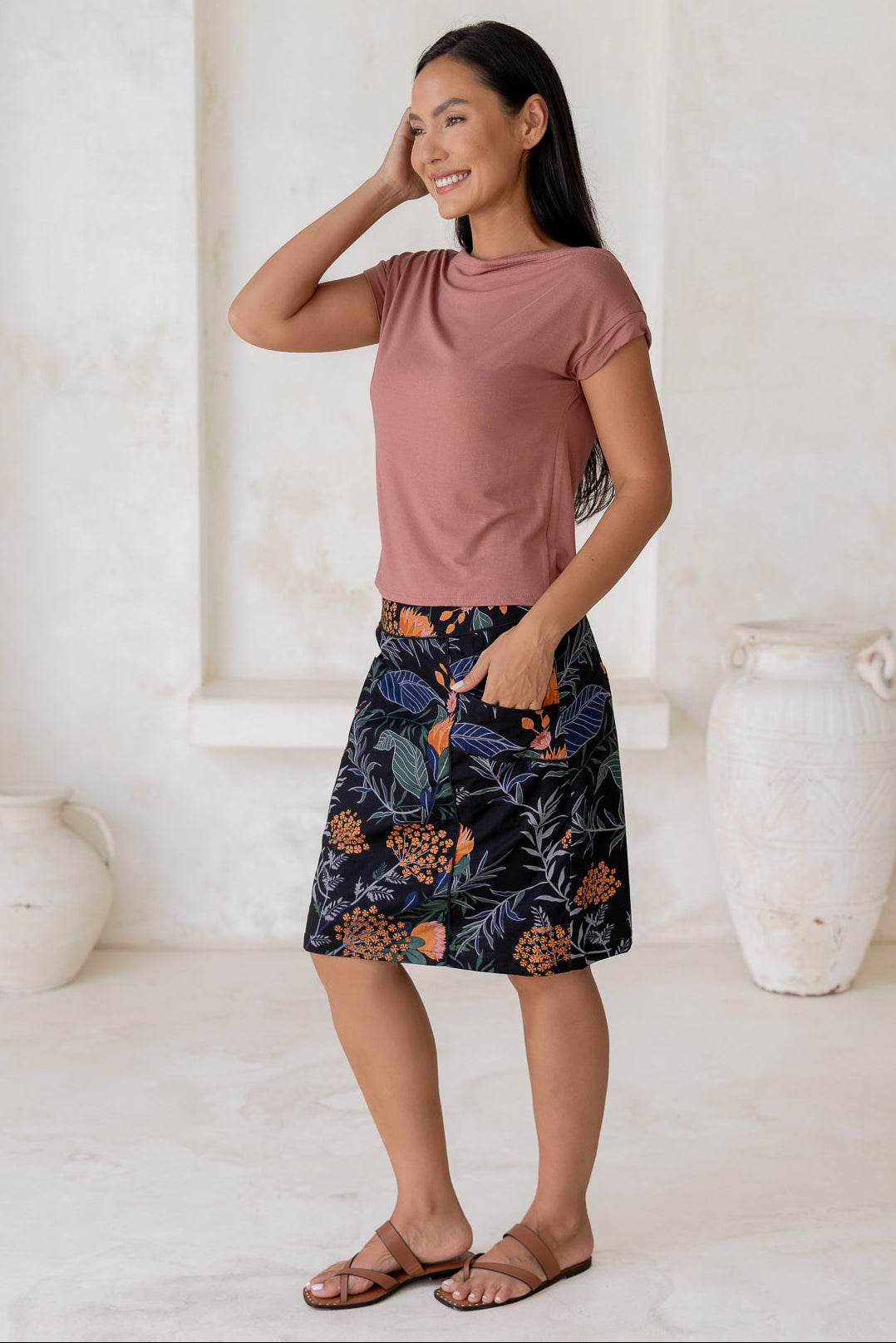 Woman posing slightly sideways wears 100% cotton A-line skirt in black based botanical print, paired with dusty rose basic top.