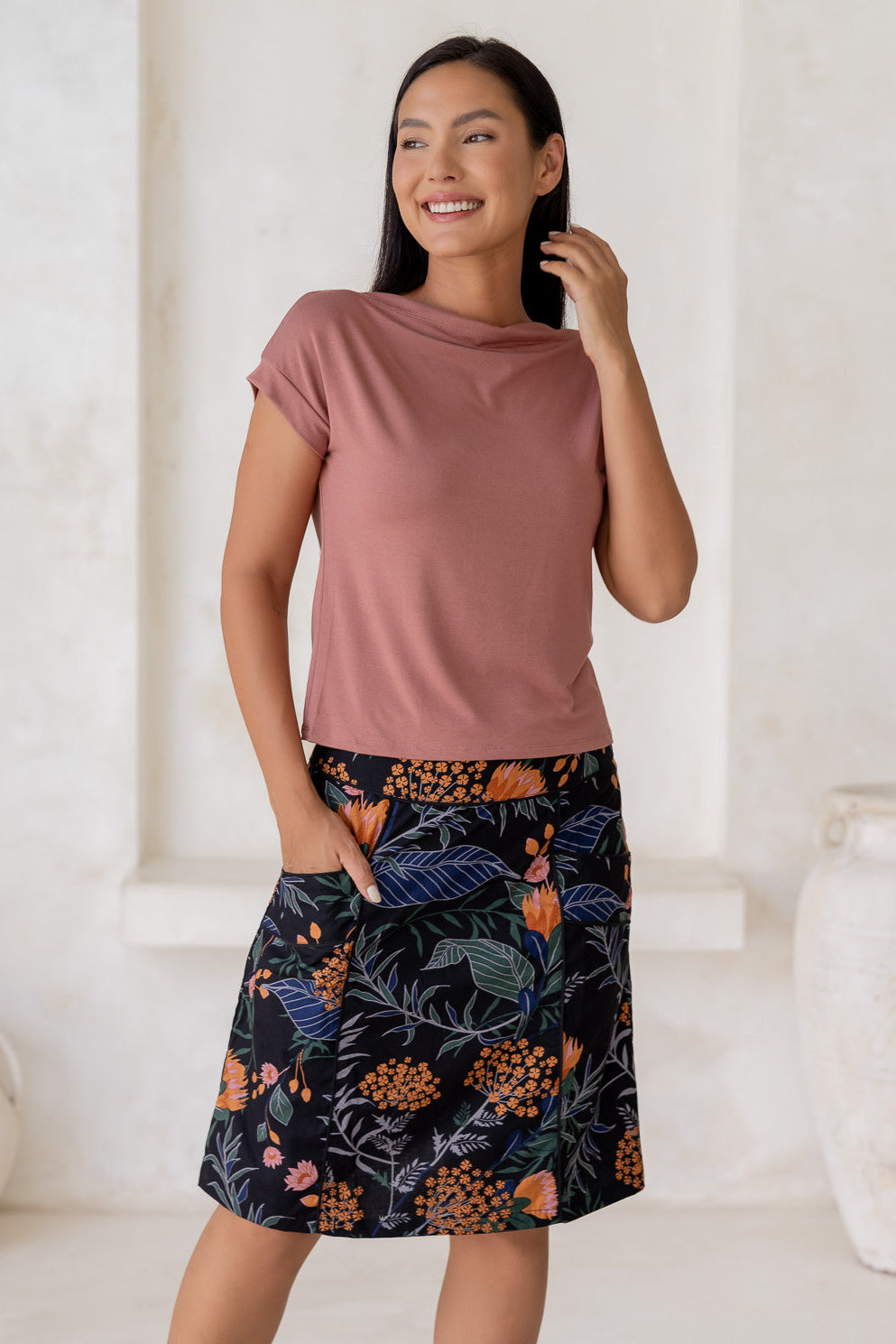 Woman stands in 100% cotton above knee length A-line skirt with piped detailing, pockets and a side zipper.