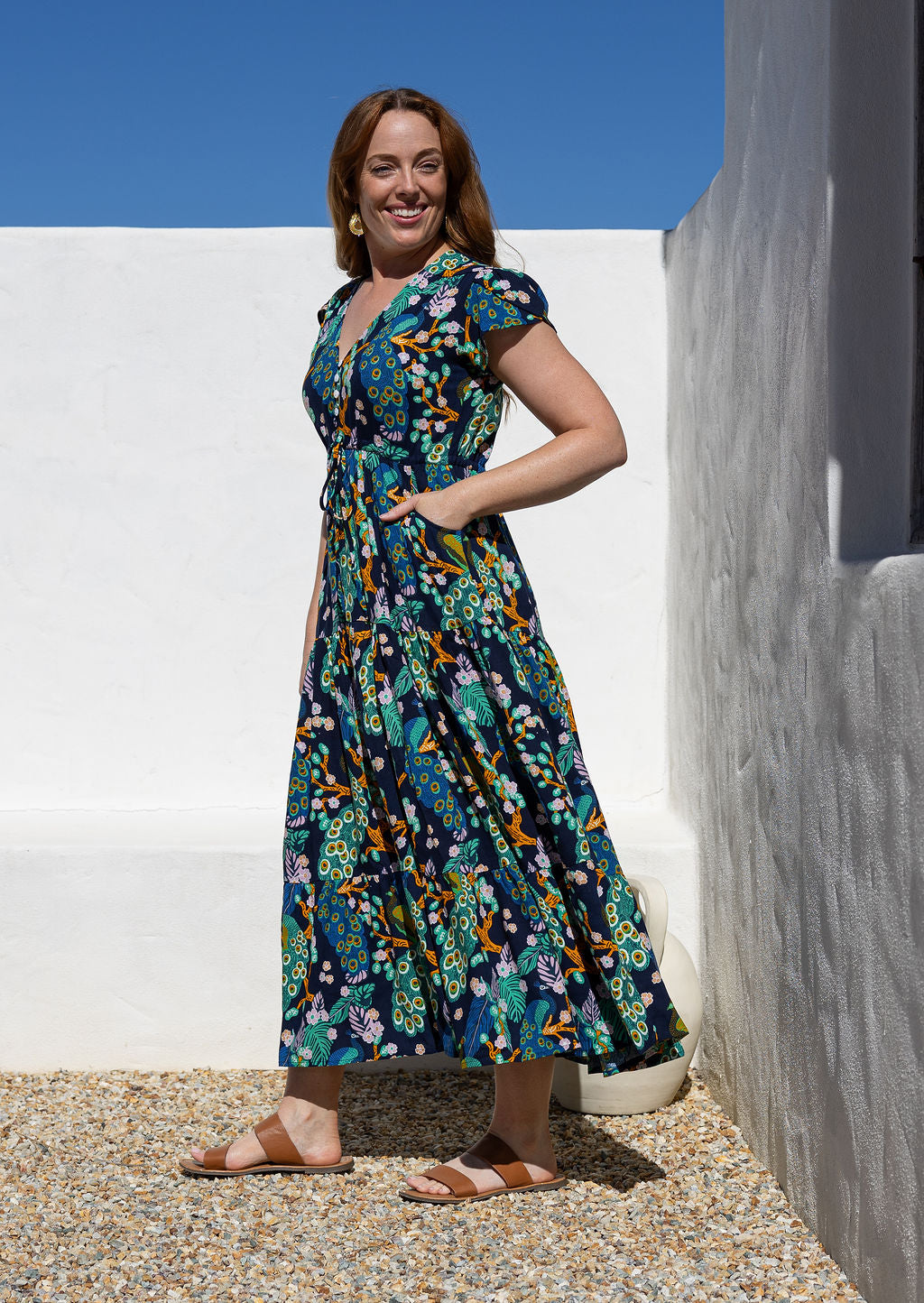 Woman standing side on wearing a rayon maxi dress with a buttoned bodice, V-neckline and cap tulip sleeves, with her hands in pockets
