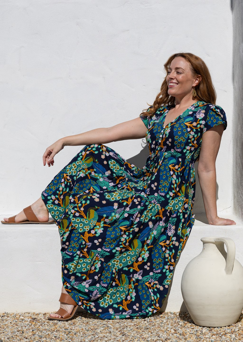 Woman wearing a short-sleeve maxi dress with a colourful peacock print in teal, blue, yellow, and orange, sitting on a white stone ledge next to a white ceramic jug.