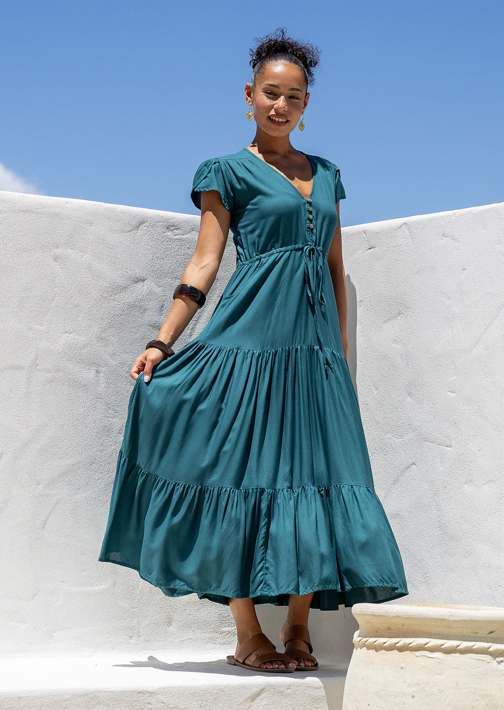 Woman in a teal maxi dress with full skirt standing against a white wall with blue sky.