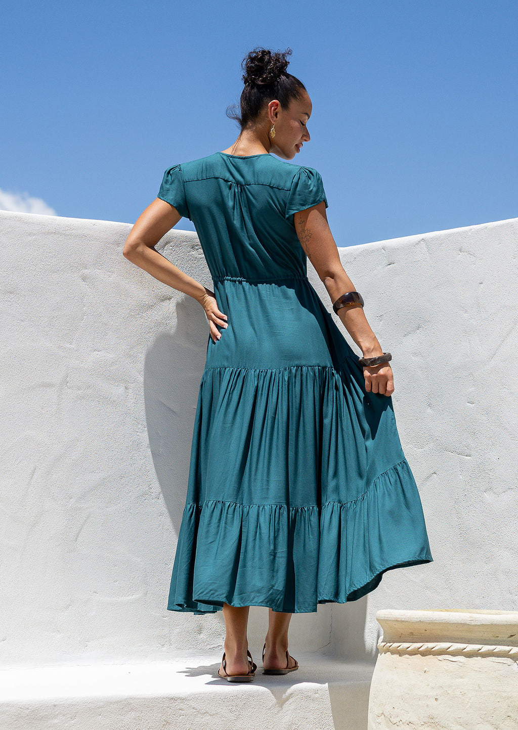 Back view of Woman in a teal dress standing against a white wall with blue sky.
