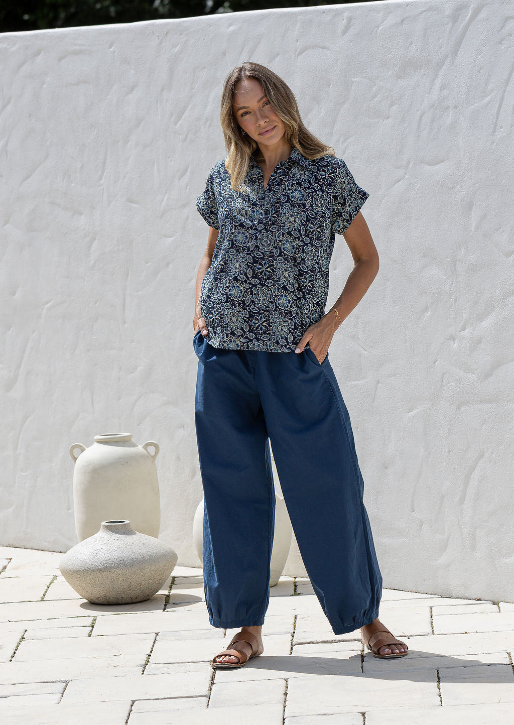 Woman wearing a navy blue based floral 100% cotton top and plain pants standing in a rustic indoor setting.