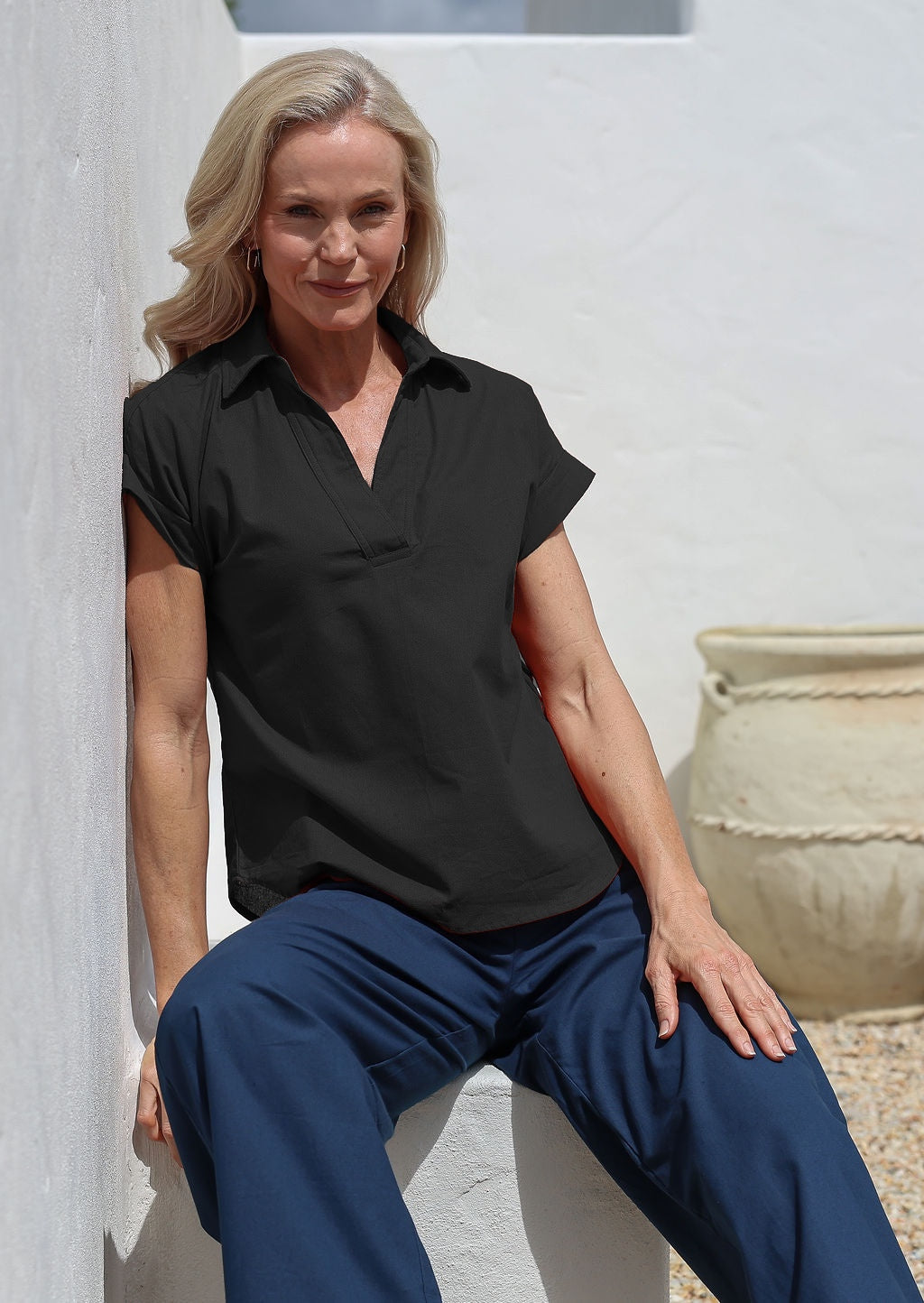Woman wearing a black cotton short sleeved collared top and blue pants sitting against a white wall.