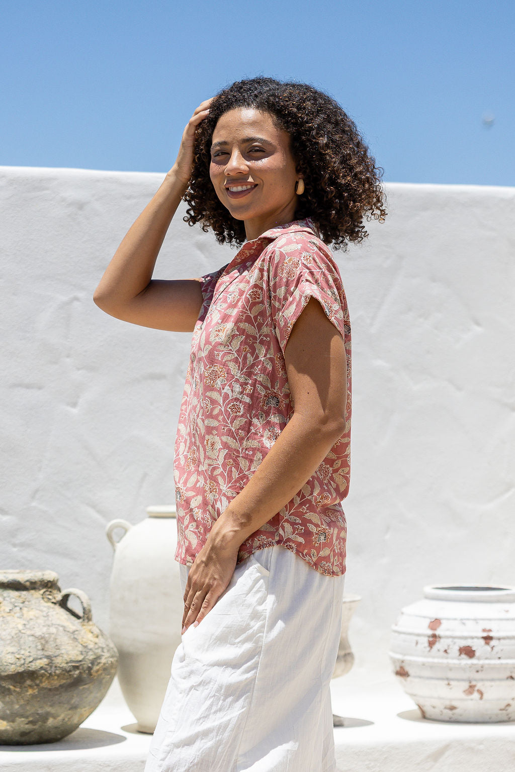 Woman stands sideways wearing a rose pink based floral cotton top with short folded cuff sleeves, collar and V neckline