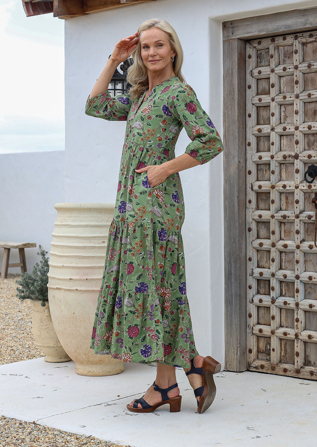 Woman posing with one hand in pocket wearing a 100% cotton green floral maxi  dress, standing in front of a wooden door.