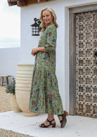 Woman in green floral, cotton maxi dress with tiered skirt, 3/4 sleeves, pockets and a mandarin collar