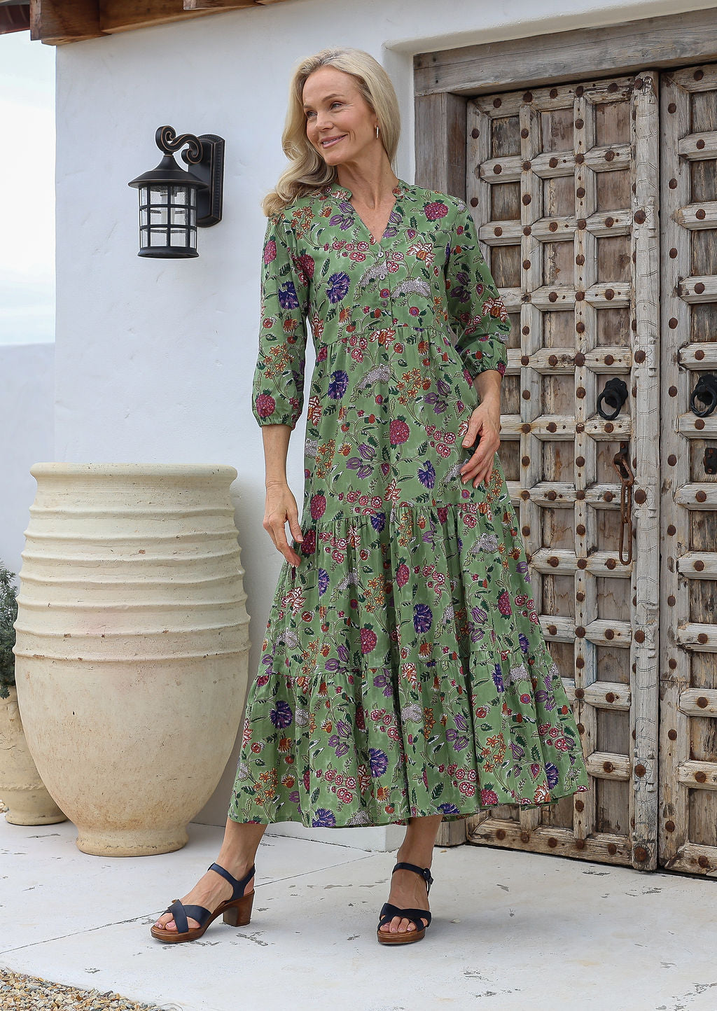 Woman wearing a green floral maxi dress with 3/4 sleeves, buttoned bodice and hands in pocket, standing in front of a rustic wooden door.