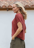 Woman smiling and standing sideways with hand in pocket wearing 100% rayon batwing top in terracotta standing in front of a white house