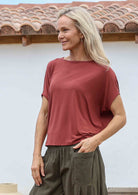 Woman with one hand in pocket wearing 100% rayon batwing top in terracotta standing in front of a white house