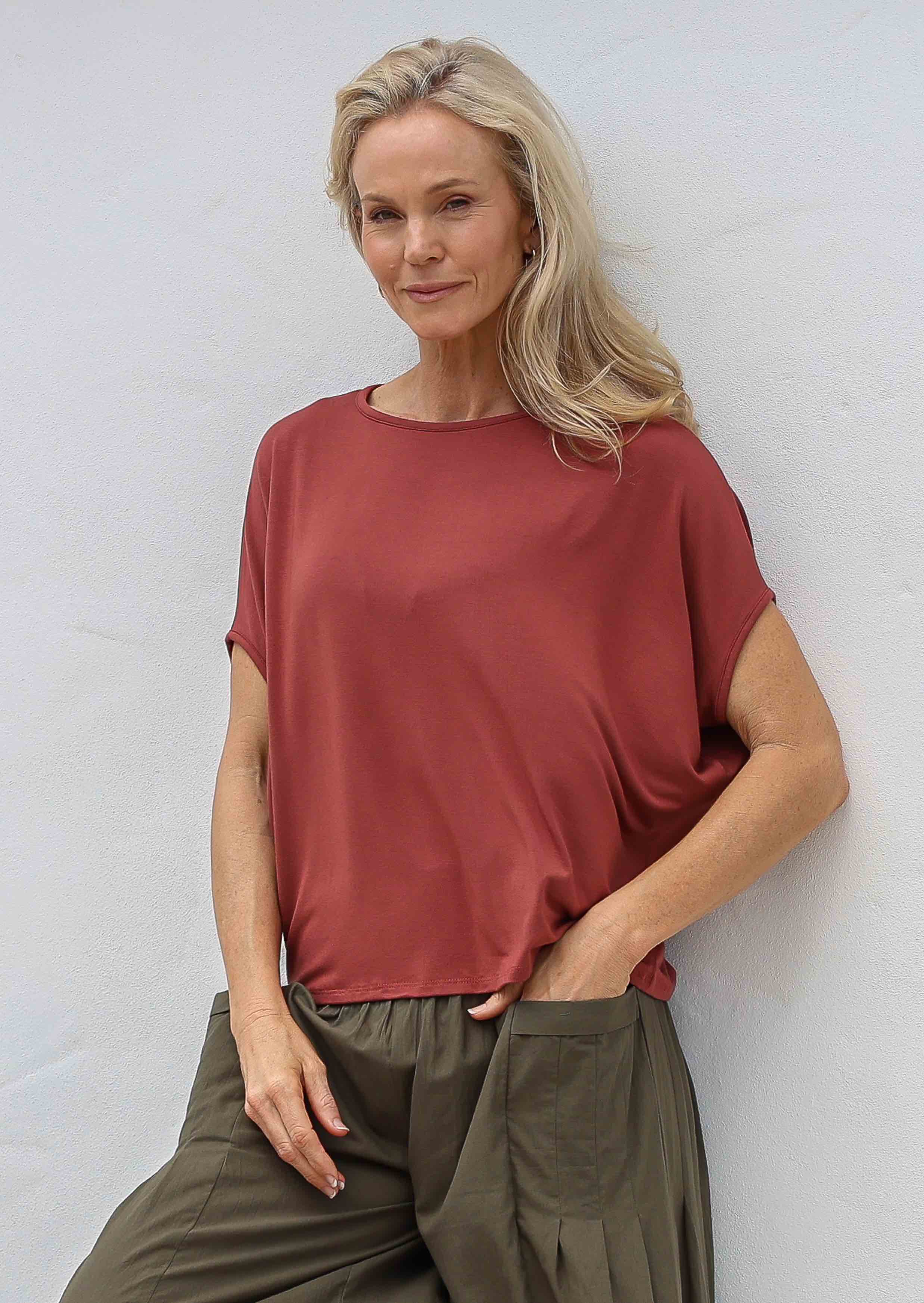 Woman with one hand in pocket wearing 100% rayon batwing top in terracotta standing in front of a white wall