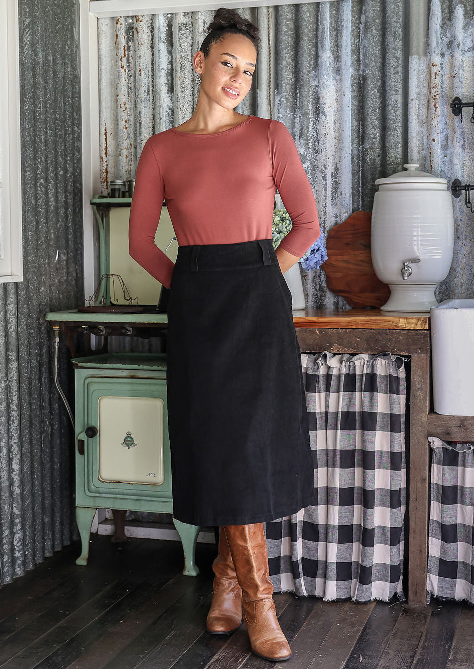 Woman in a rustic setting wearing a black cotton corduroy A-line midi length skirt and tan boots.