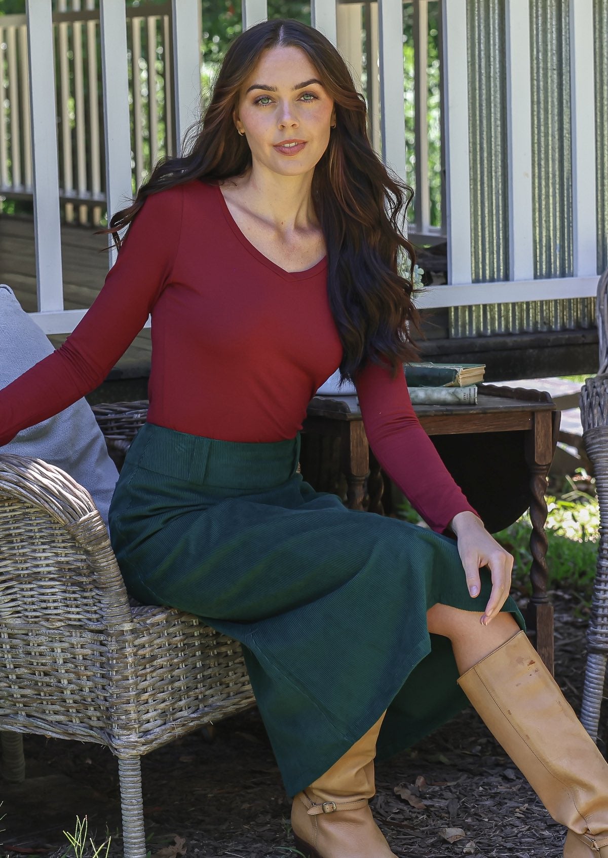 Woman sitting outdoors wearing a maroon long sleeve top and green cotton corduroy skirt with tan boots.