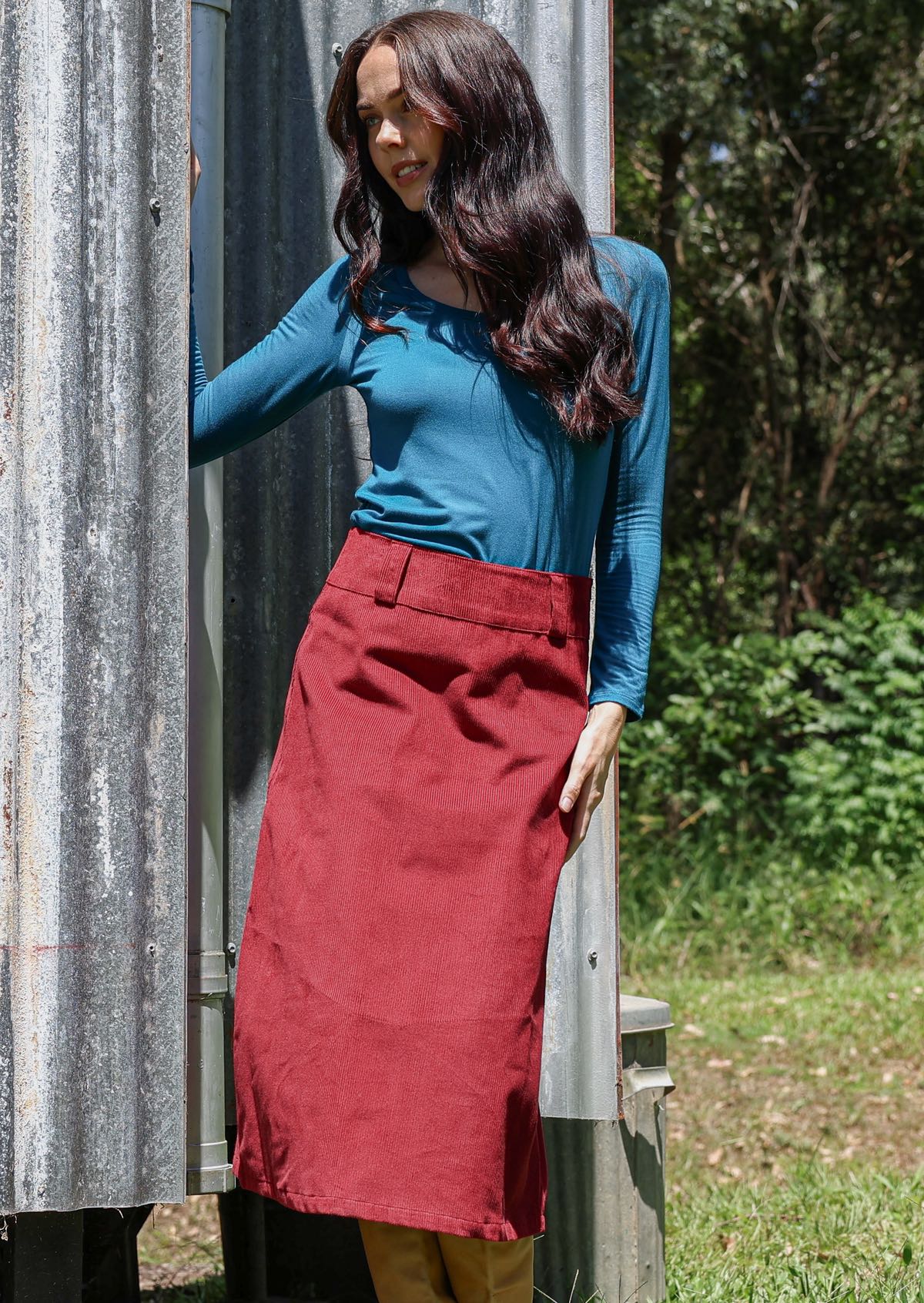 Woman in a teal top and red cotton corduroy midi length skirt standing next to a corrugated metal wall outdoors.