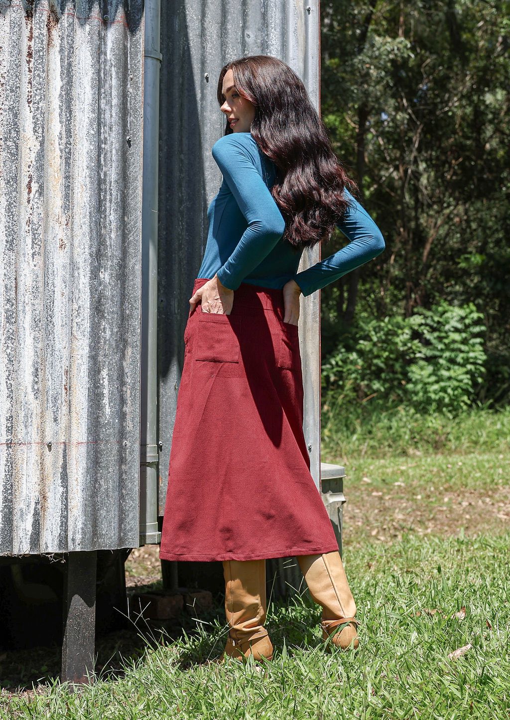 Woman with her back turned wearing a teal top and red cotton corduroy A-line midi length skirt with her hands in the back pockets
