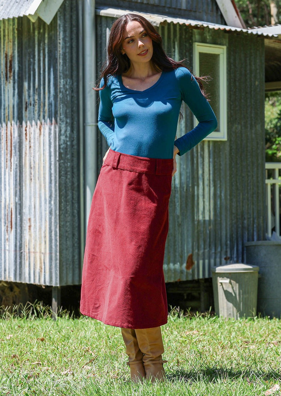 Woman wearing a cabernet red cotton corduroy A-line midi length skirt with belt loops, paired with a teal top and tan boots