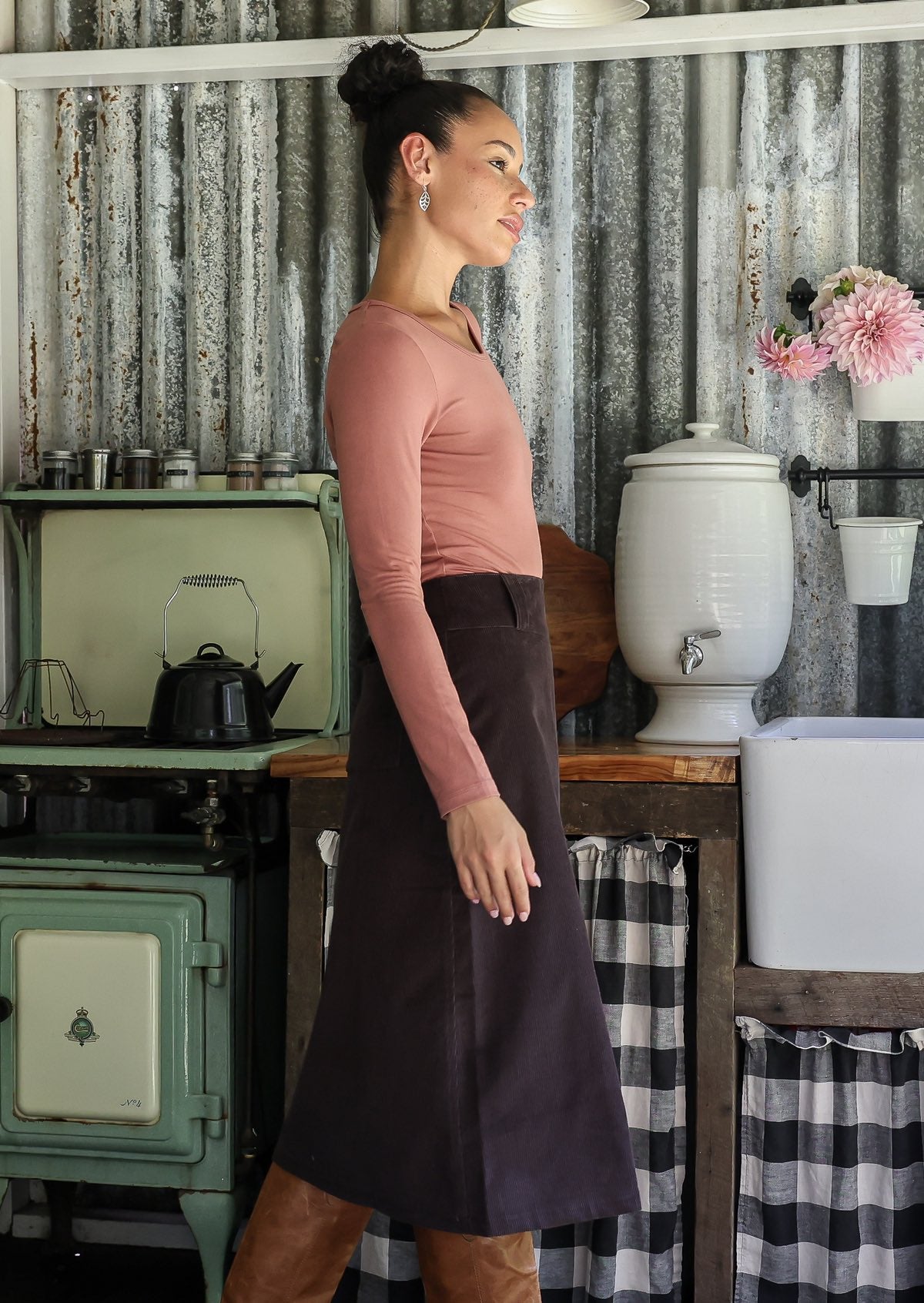 Woman standing side on wearing a chocolate brown cotton corduroy A-line midi length skirt paired with a rose pink top