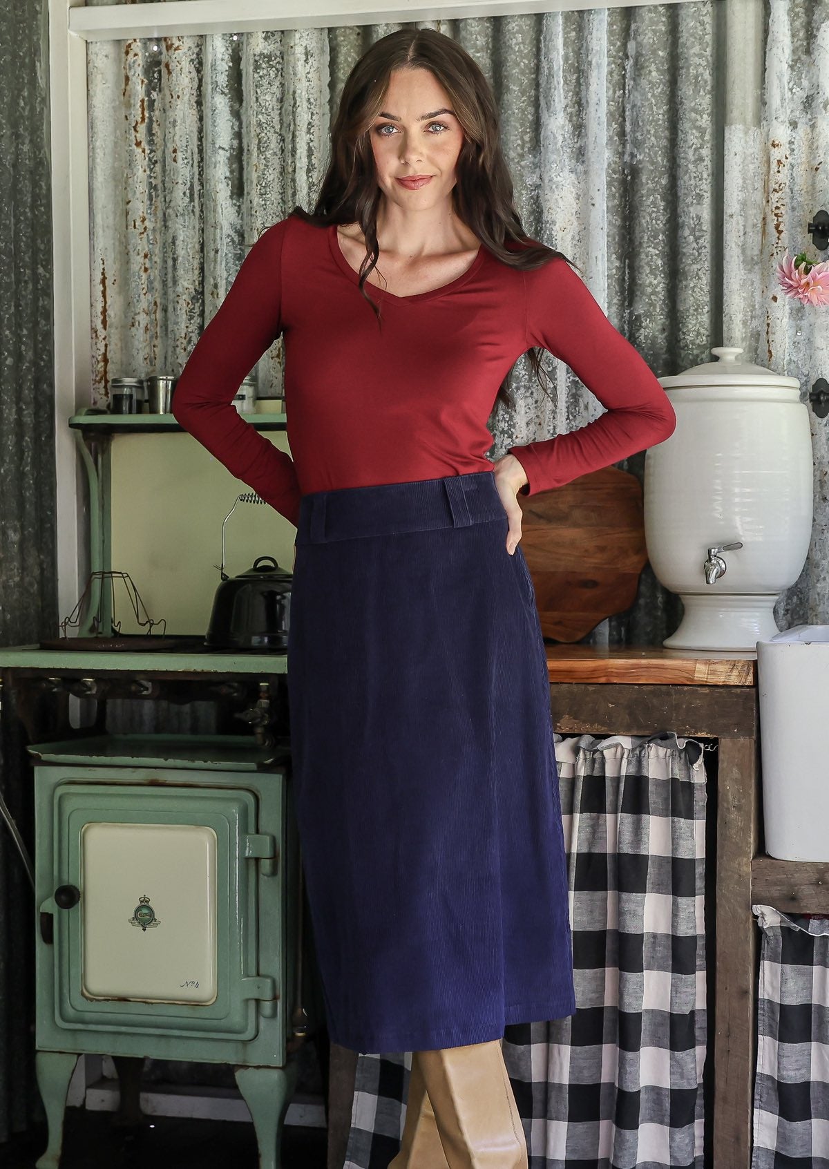 Woman in a maroon top and navy midi length cotton corduroy skirt standing in a rustic room
