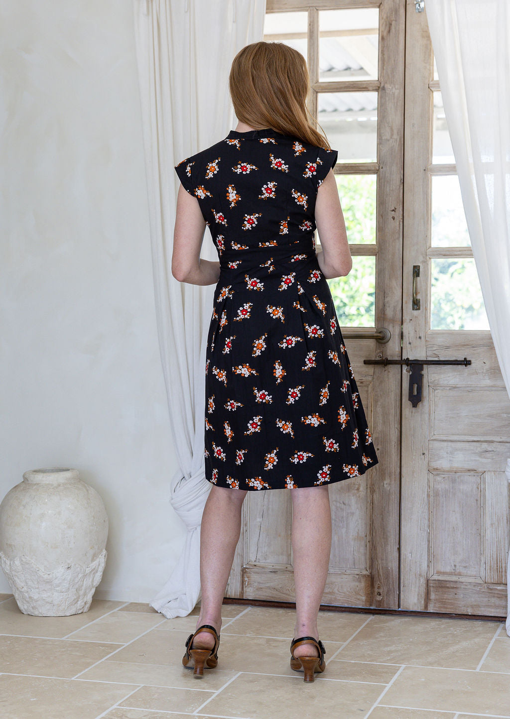 Woman stands backwards wearing 100% cotton retro style knee length dress with box pleats, cap sleeves and V neckline.