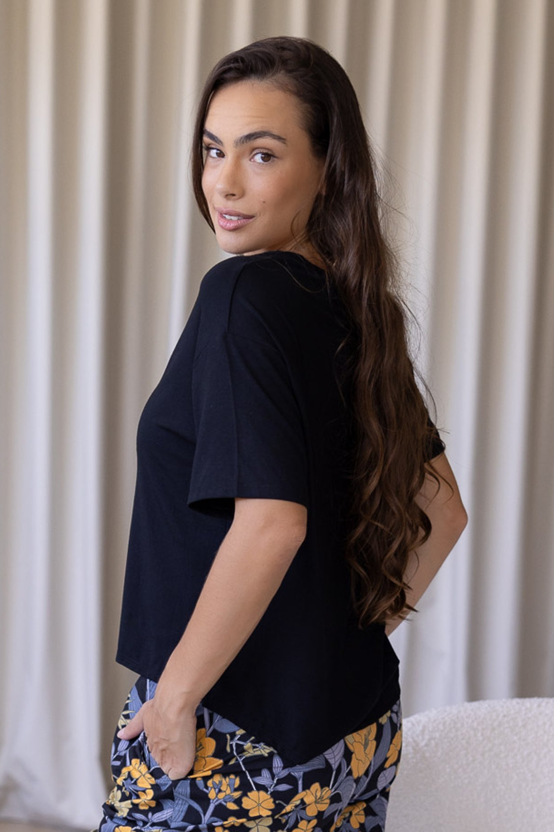 Woman side on wearing a black relaxed fit stretch rayon top with short sleeves that is longer at the back